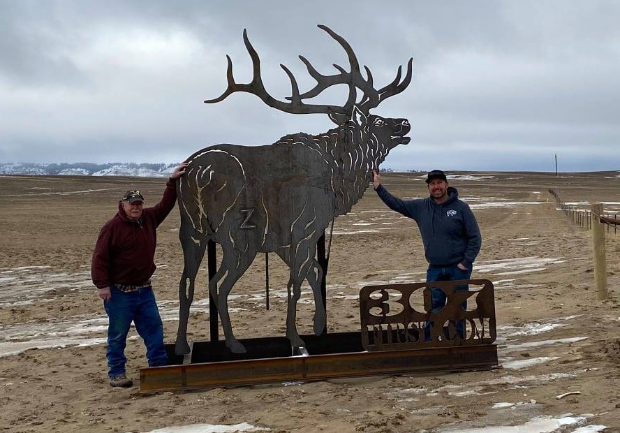 Two men stand next to elk metal silhouette